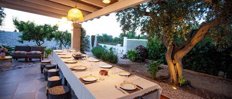 Ein Essbereich im Freien mit einem langen Tisch, gedeckt für eine Mahlzeit, umgeben von hölzernen Hocker und unter einer Holzpergola mit hängenden Lichtern.