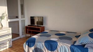 Une chambre à coucher avec un lit, une commode, un téléviseur posé sur un meuble et un vase rempli de fleurs sur la commode.