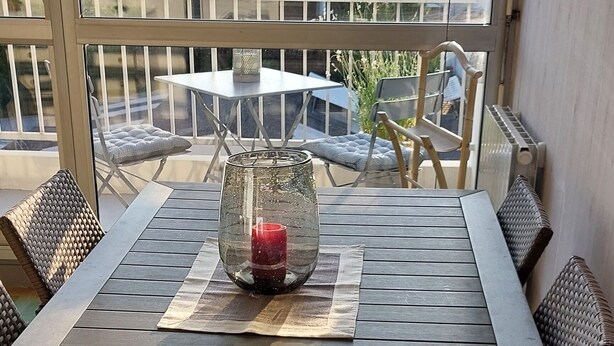 Une table en bois, sur laquelle sont posés un vase en verre et une bougie, est placée sur un balcon offrant une vue sur le paysage urbain.