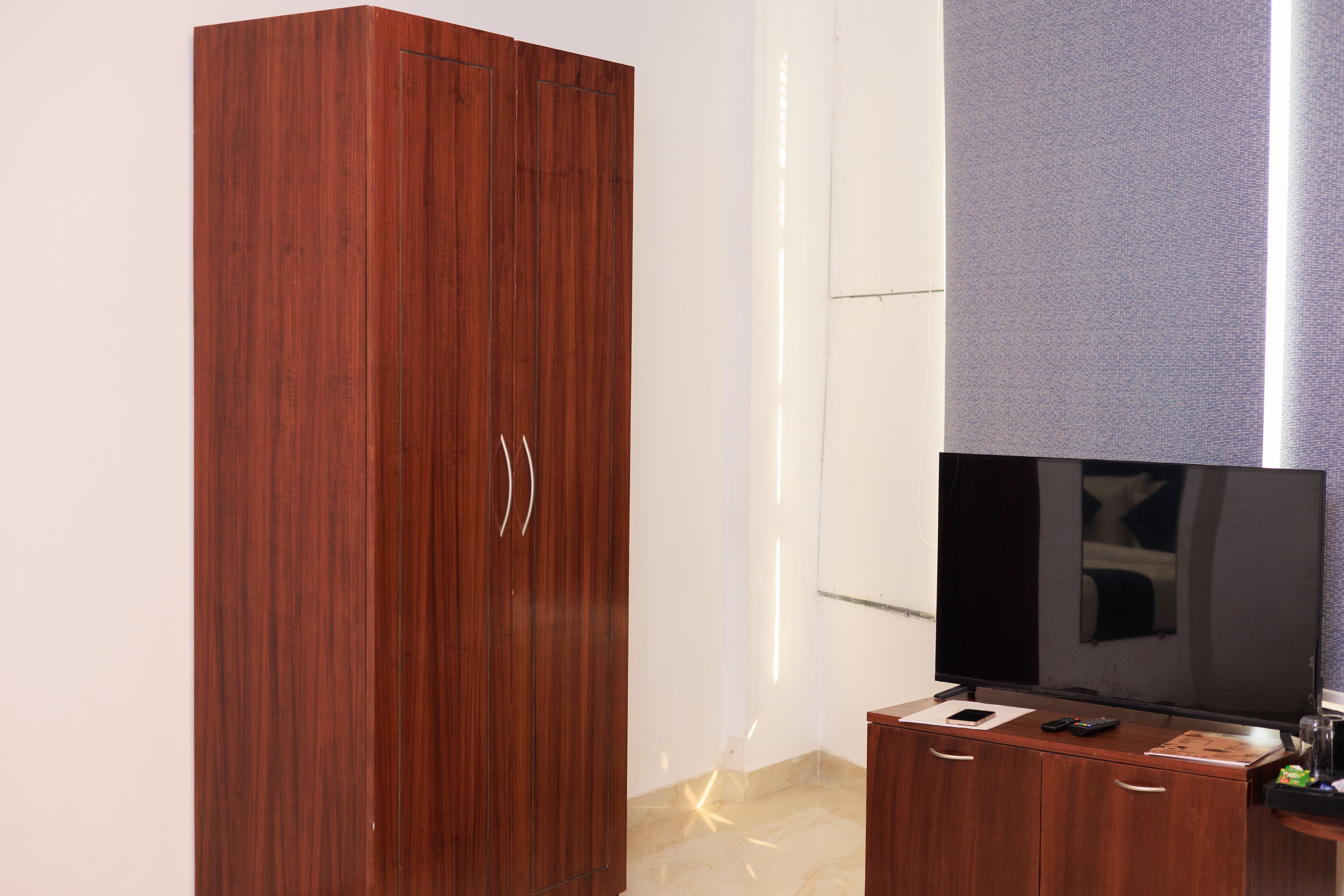 A room with a wooden wardrobe, a flat-screen TV on a wooden stand, and a small table with a remote control.