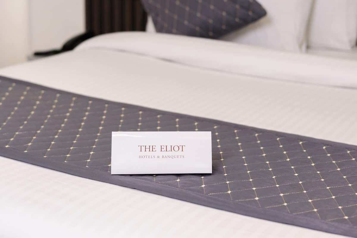 Cama arrumada com uma placa indicando "THE ELIOT HOTELS & BANQUETS" sobre um edredom estampado em tons escuros.