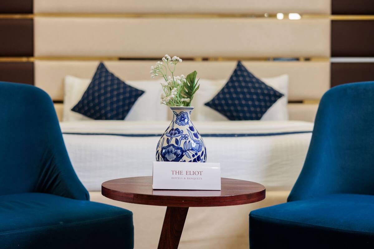 Quarto de hotel com uma cama, duas cadeiras azuis, uma mesa de madeira com um aviso e um vaso azul e branco com flores.