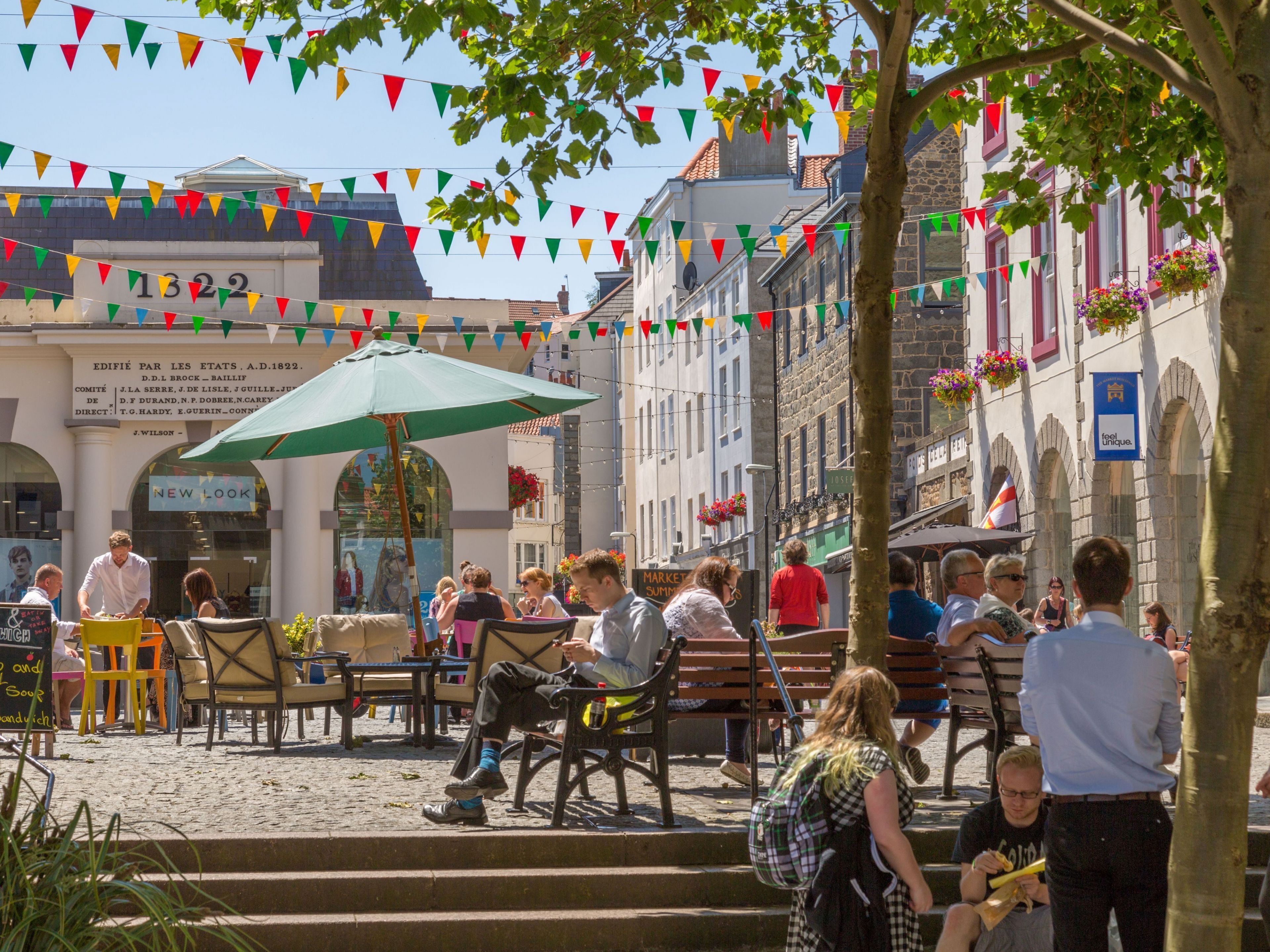 Un café animé en plein air, avec des tables et des chaises, entouré de bâtiments historiques et de drapeaux aux couleurs vives.