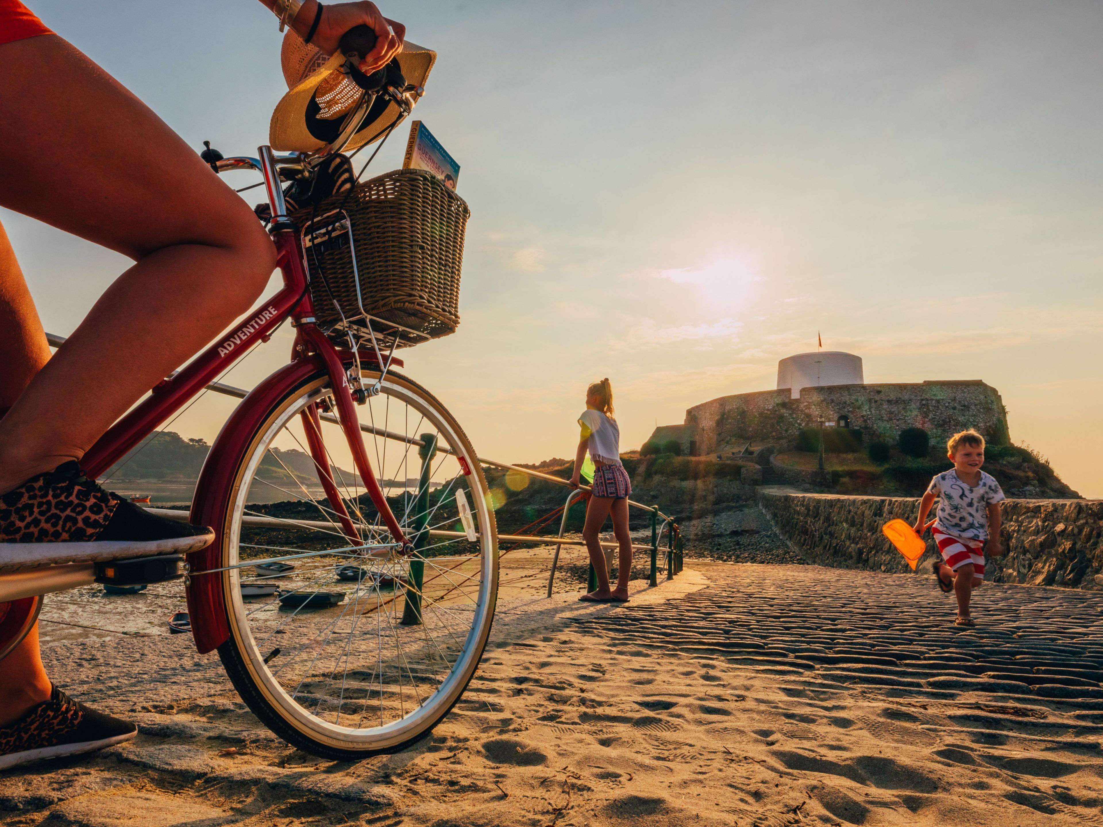 En strandscene med en cykel, en person i leopardmønstrede sko, og et barn med et strandlegetøj.