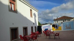 An outdoor patio with red chairs and tables, a white building with windows, and a mural on the adjacent wall. - Albergue Miraiso (Arzua)