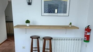 A kitchen with a white countertop, two wooden stools, a framed picture, a small potted plant, a wall-mounted light, and a fire extinguisher. - Albergue Miraiso (Arzua)