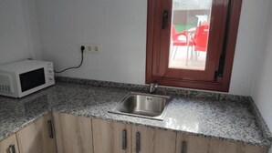 A kitchen with a granite countertop, a stainless steel sink, wooden cabinets, a microwave, and a window with a view of outdoor chairs. - Albergue Miraiso (Arzua)