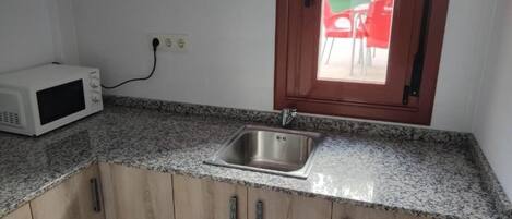 Una cocina con encimera de granito, pileta de acero inoxidable, muebles de madera, un microondas y una ventana con vistas a sillas exteriores.