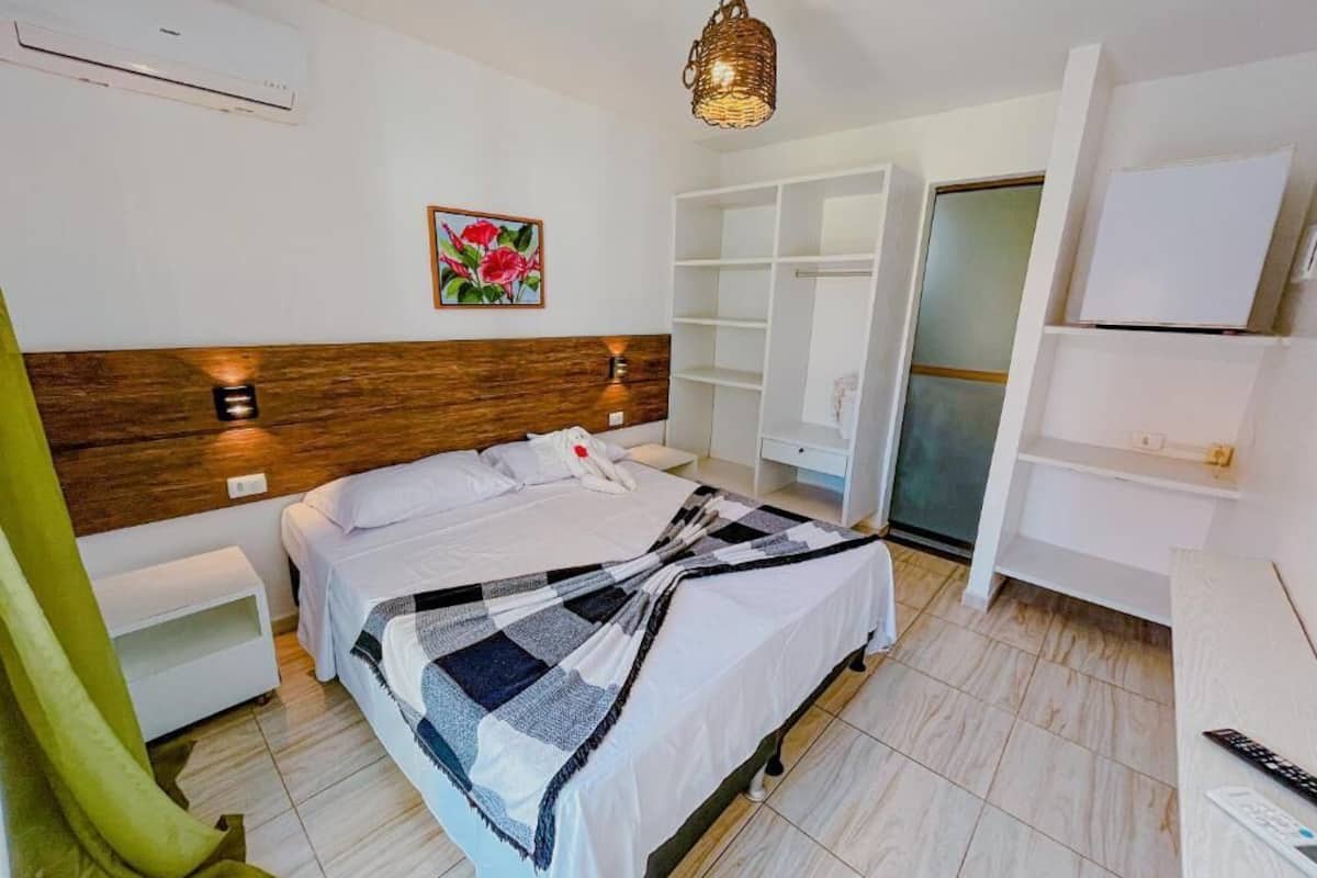 Un dormitorio con cabecera de madera, una cama con manta a cuadros, una mesita de noche, una silla verde, un armario y una lámpara colgante.