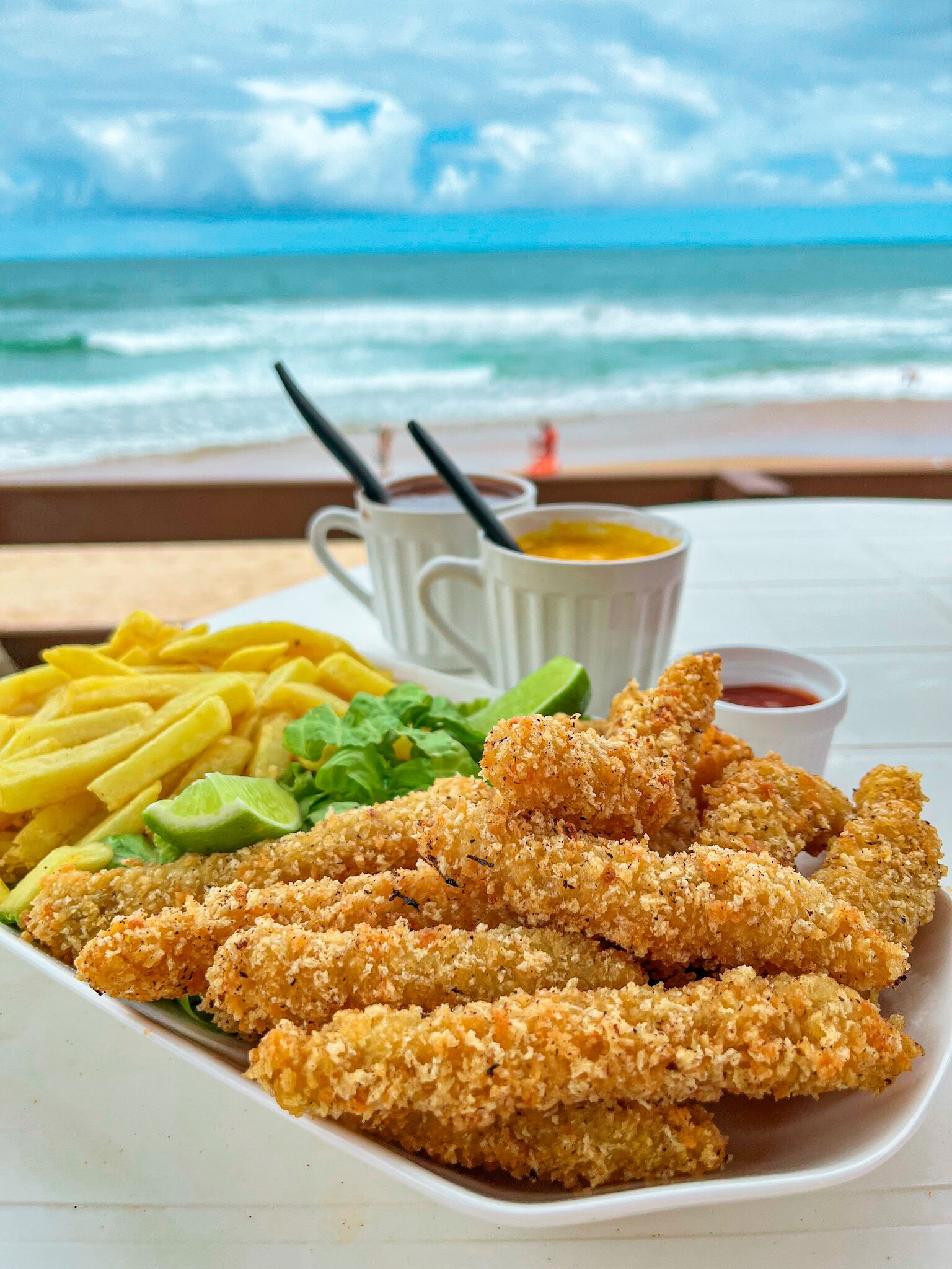 A plate of food with fries, avocado, and breaded chicken, with a view of the ocean and beach in the background.