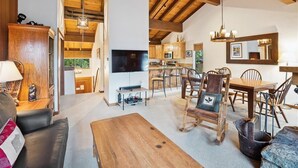 Un salón amplio con techo de madera, televisor, mesa de comedor con sillas y cocina al fondo.