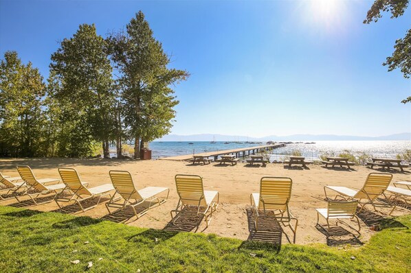 A beachside setting with sun loungers and picnic tables arranged on the sand, overlooking a calm sea with boats and a pier in the distance.