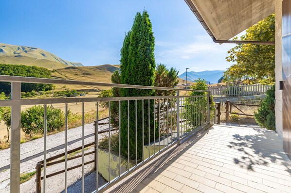 Een balkon met een metalen hekwerk dat uitkijkt over een glooiend landschap met een hoge altijdgroene boom en een heldere lucht.