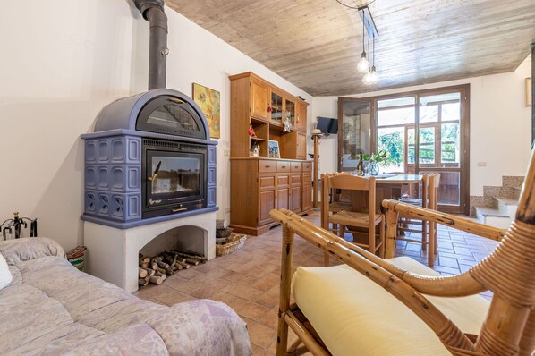 A cozy living room with a blue wood-burning stove, a wooden cabinet, and a dining area with wicker chairs. - Italian Experience - Casa TraMonti (Bolognola)