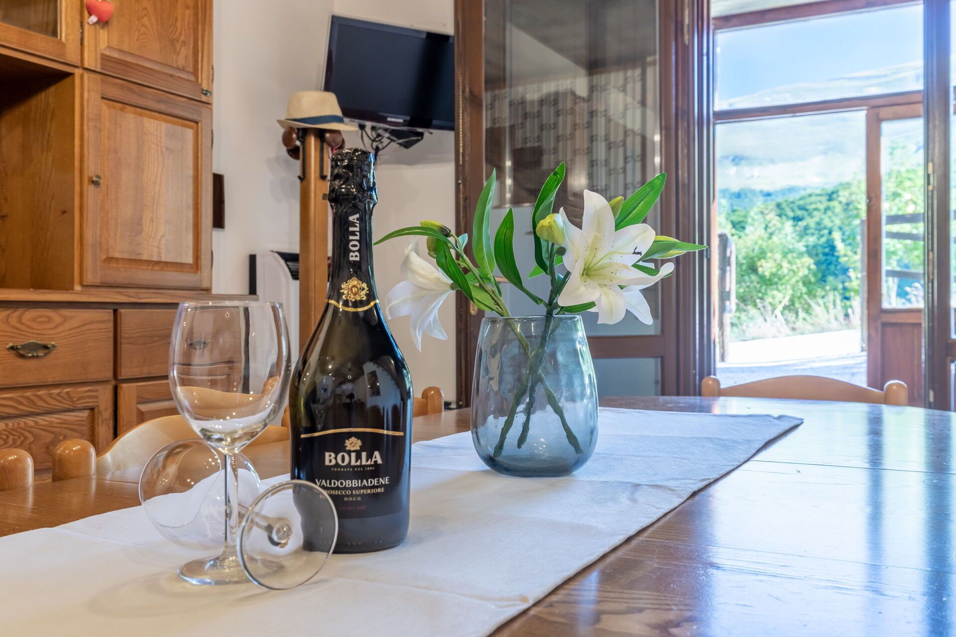 A bottle of Bolla Valdobbiadene wine, a glass wine glass, and a vase with white lilies on a wooden table.