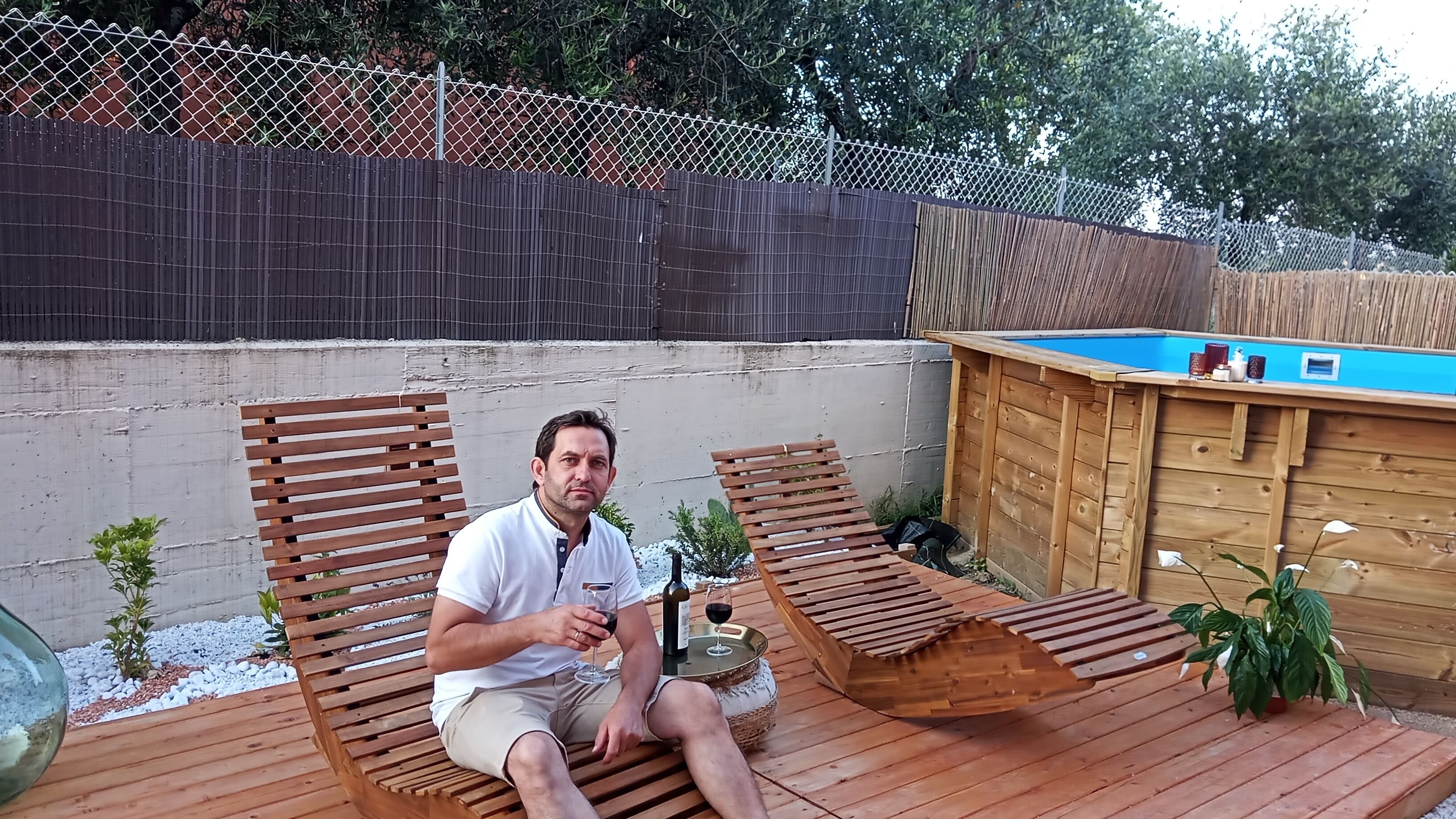 Un hombre sentado en una terraza de madera, con una piscina al fondo.