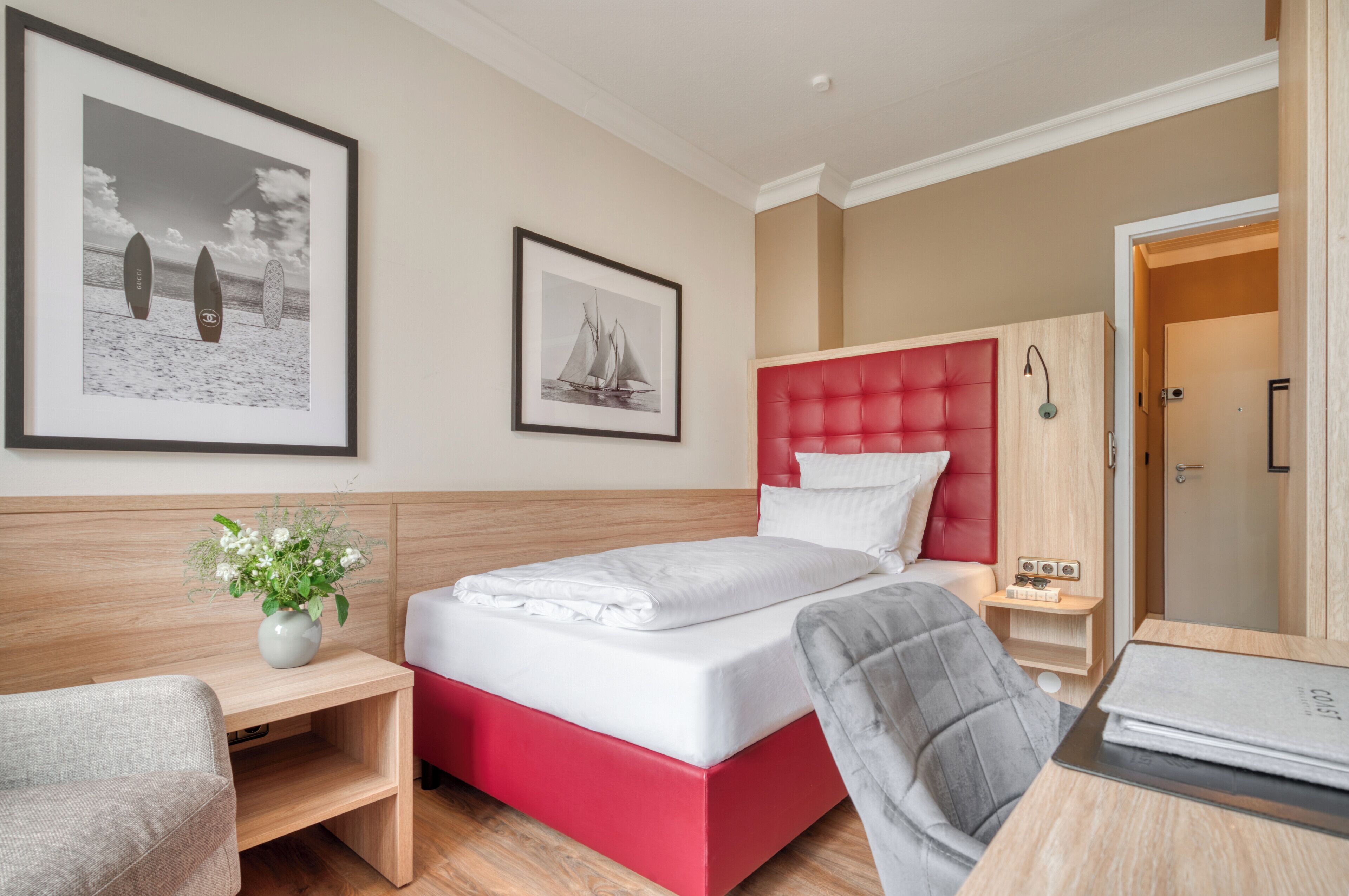A hotel room with a bed, a desk, a chair, a wardrobe, and two framed pictures on the wall.