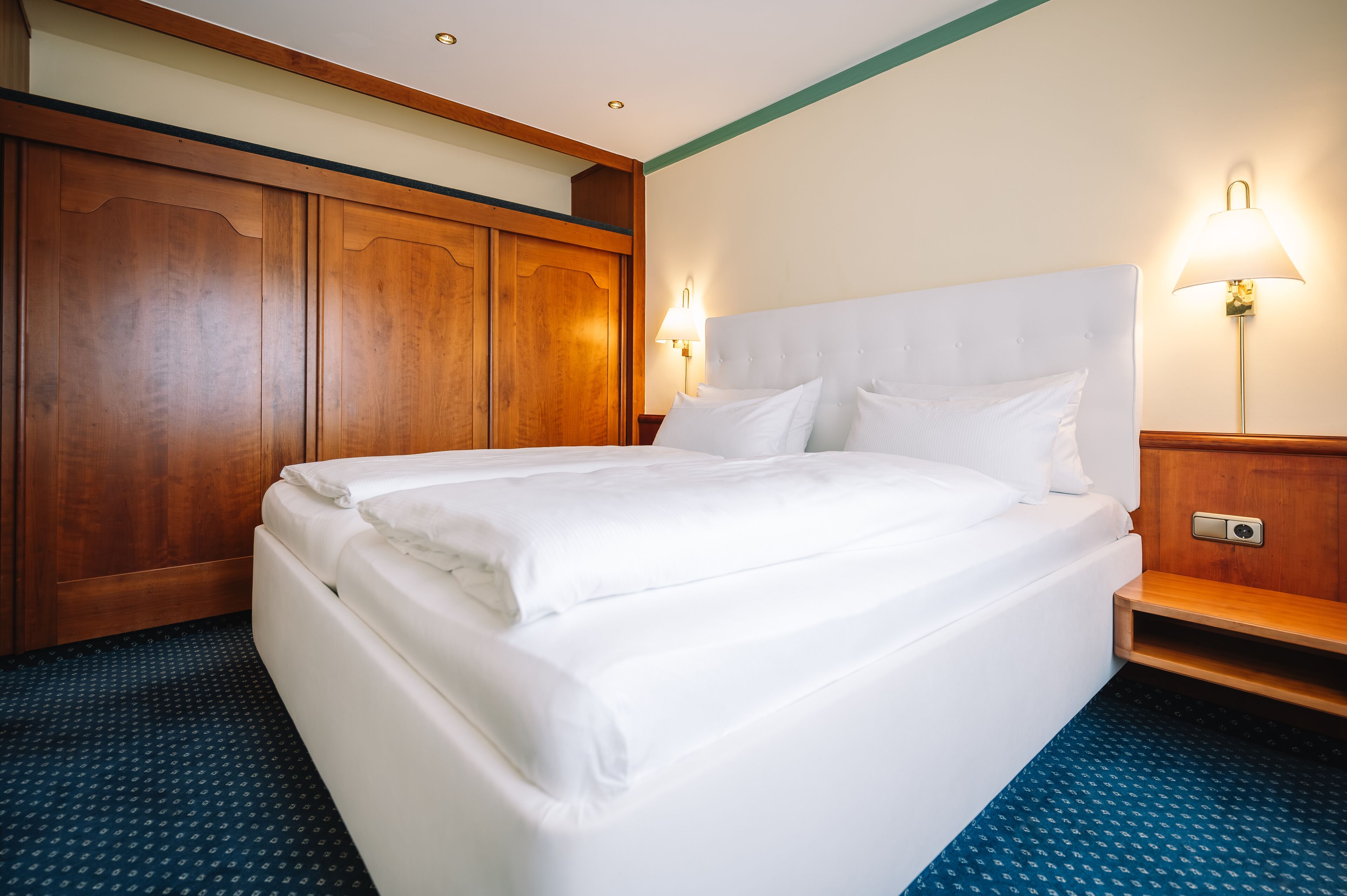 A neatly made bed in a hotel room with wooden paneling and a blue carpet.