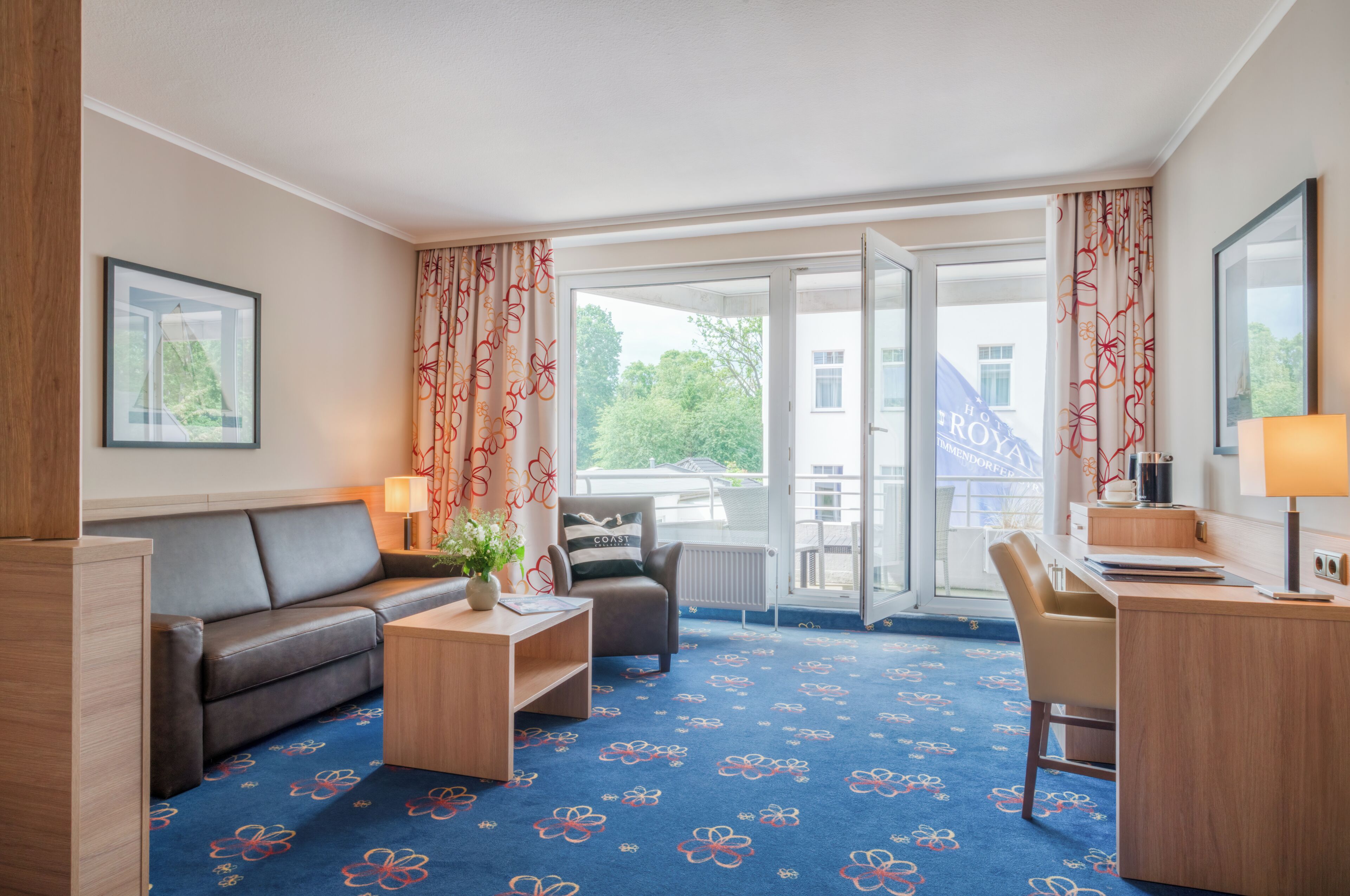 A hotel room with a sofa, a small table, a chair, a desk, and a view of the outdoors through the glass door.