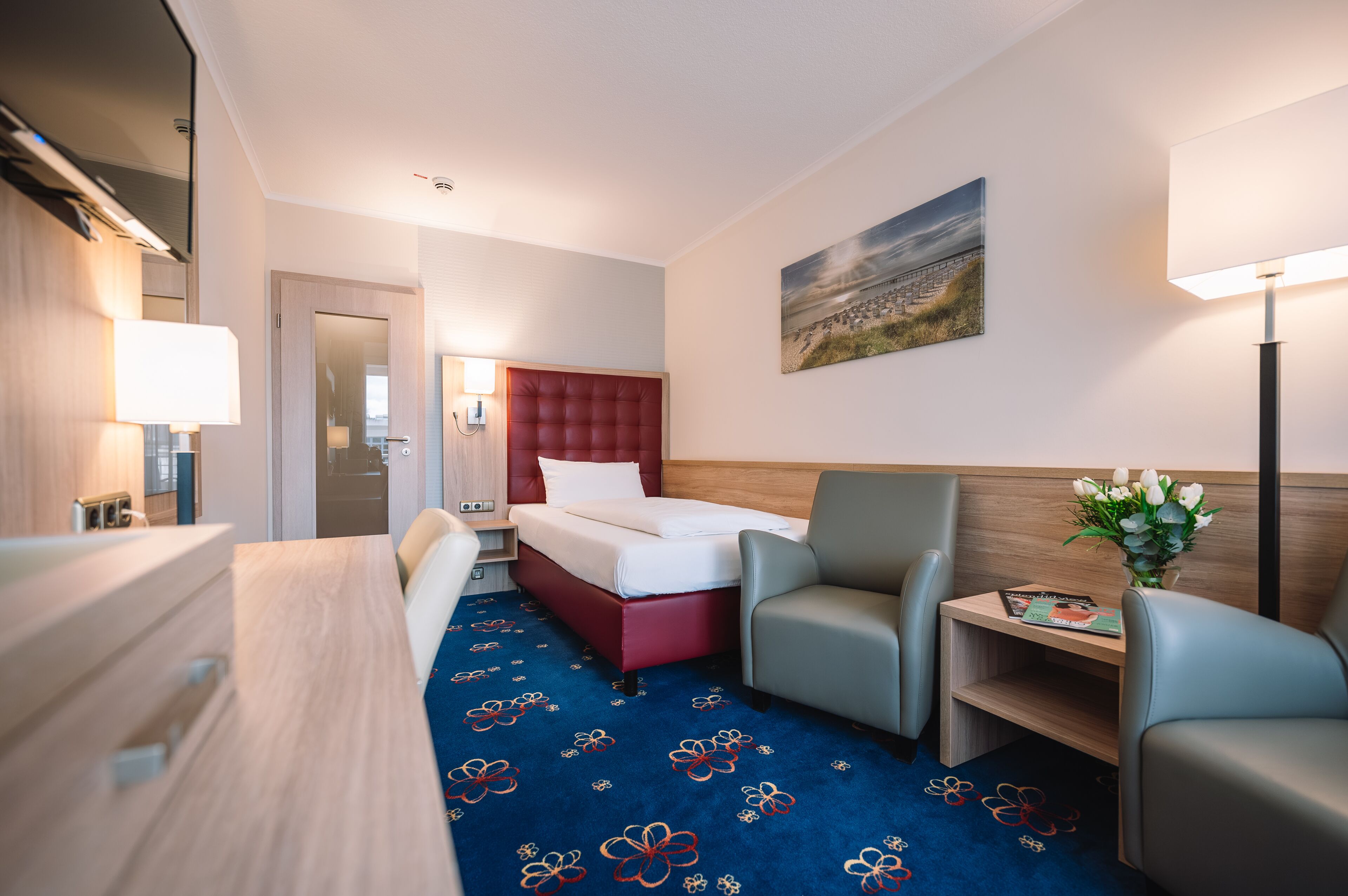 A hotel room with a bed, a desk, a chair, a lamp, and a painting on the wall.
