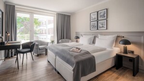 A modern hotel room with a large bed, a desk, and a chair. There are curtains, a nightstand with a lamp, and framed pictures on the wall. - Hotel Royal - Timmendorfer Strand (Timmendorfer Strand)