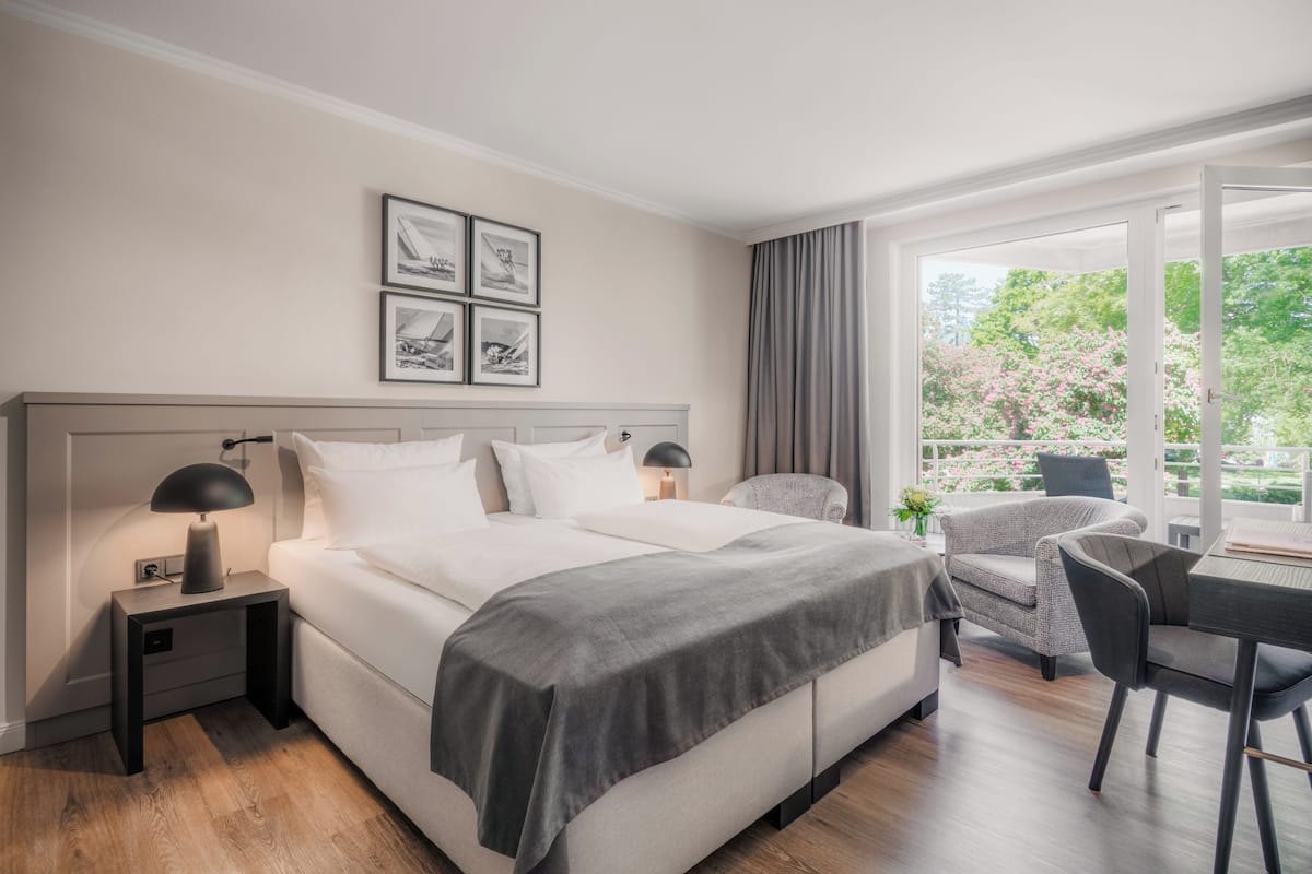 A modern hotel room with a large bed, bedside tables, a desk, and a chair. There are framed pictures on the wall and a view of greenery outside.
