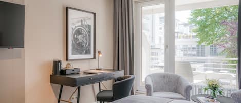 Ett hotellrum med en säng, ett skrivbord med en stol, en tv, en inramad tavla och ett fönster med utsikt över byggnader och träd.