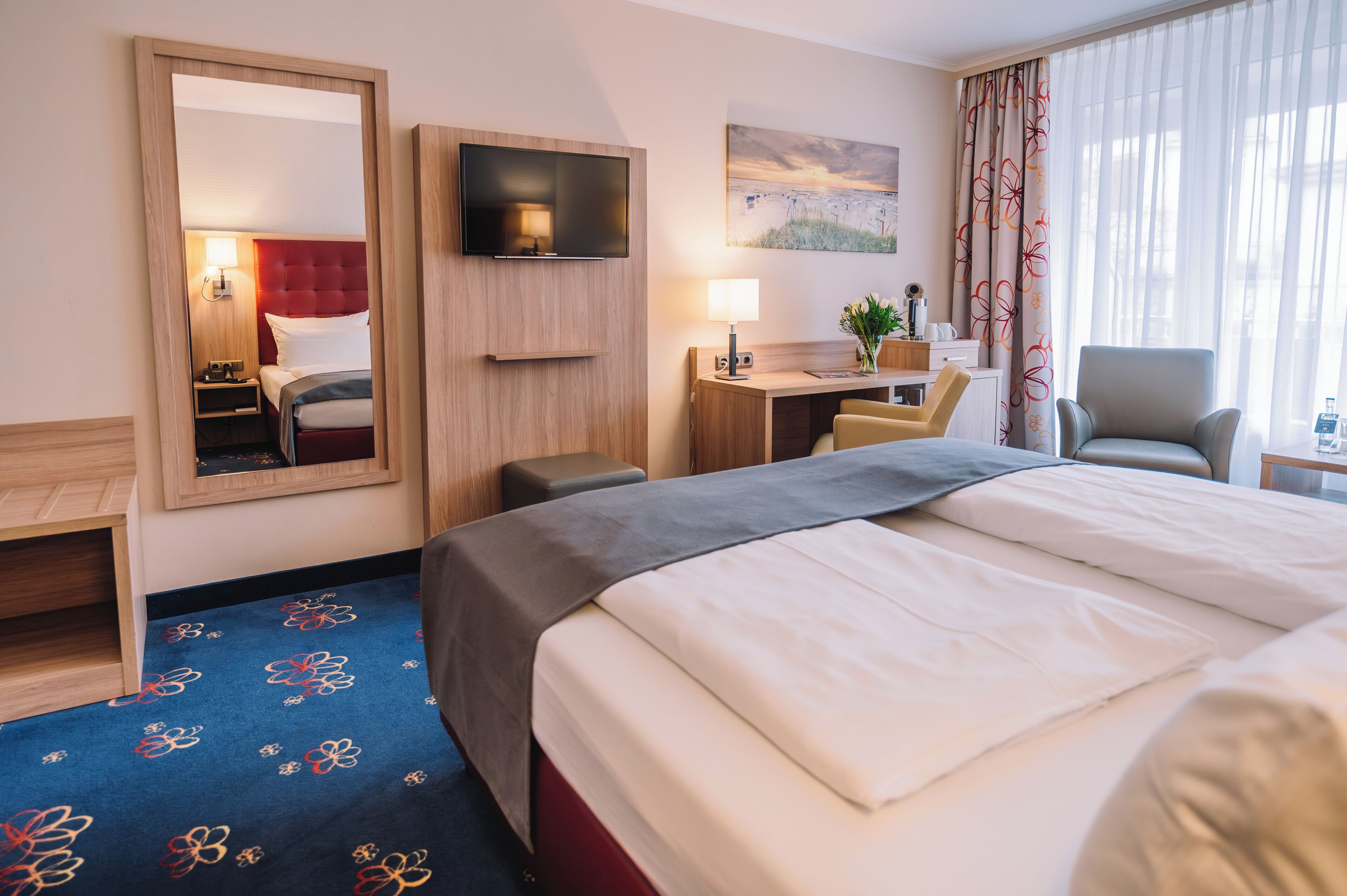 A hotel room with a large bed, a desk with a chair, a TV mounted on the wall, and a window with curtains.