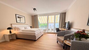 A hotel room with a bed, a sofa, a TV, a table with a vase of flowers, and a balcony with curtains. - Hotel Royal - Timmendorfer Strand (Timmendorfer Strand)