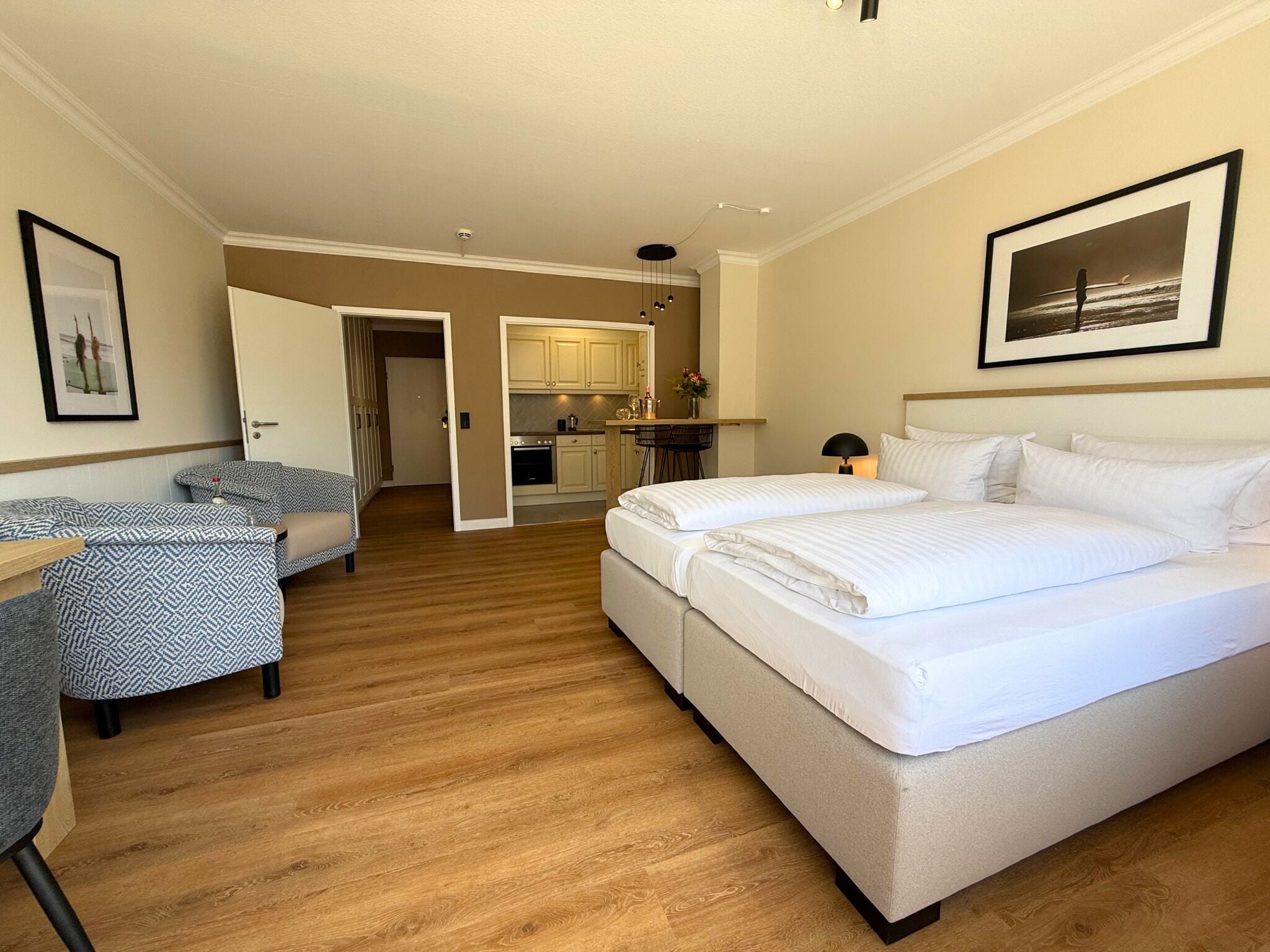 A hotel room with a bed, a desk, a chair, a kitchenette, and a door leading to another room.