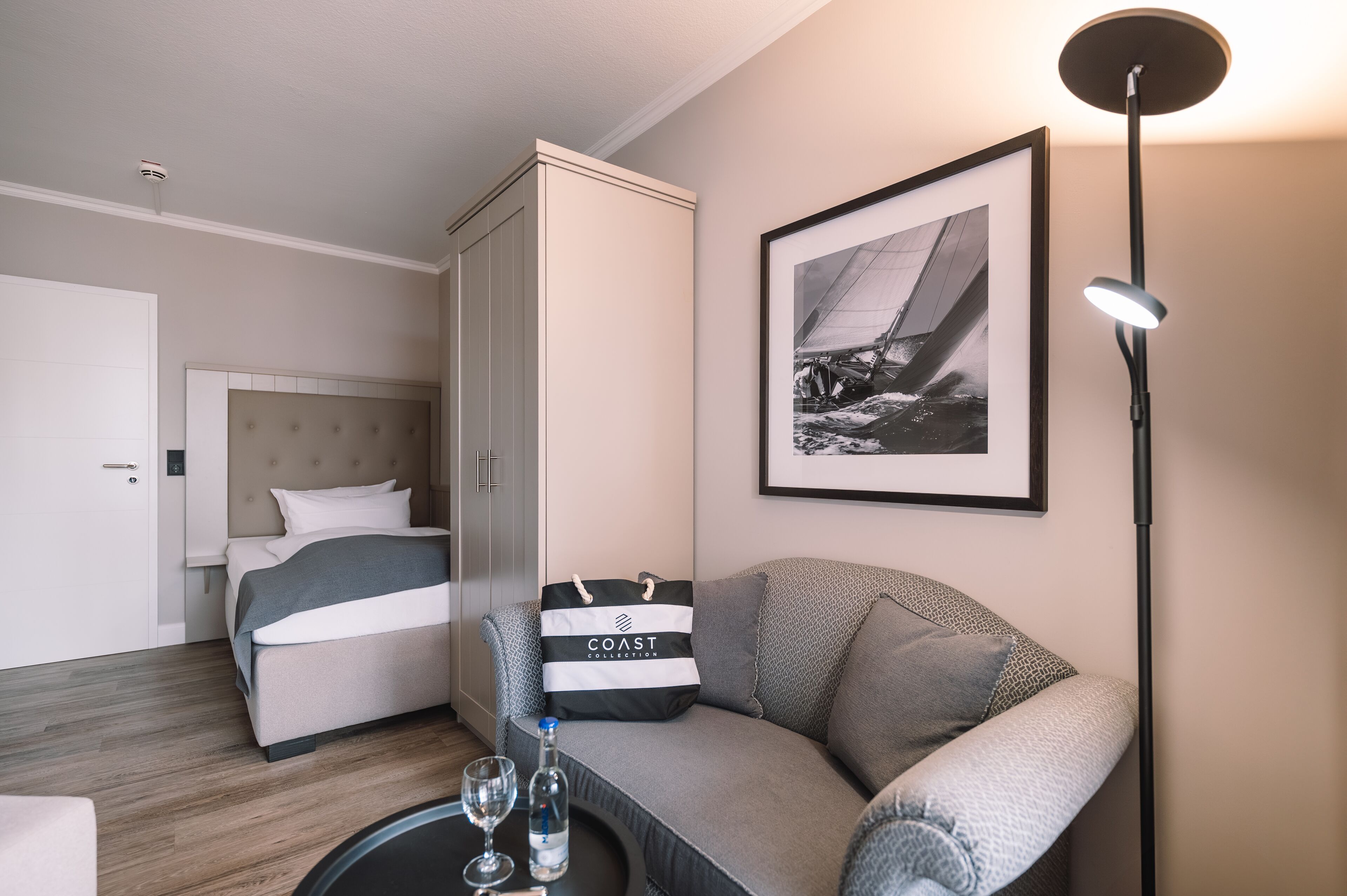 Et hotelværelse med en seng, en sofa, et billede i ramme på væggen, en gulvlampe og en taske på sofaen.