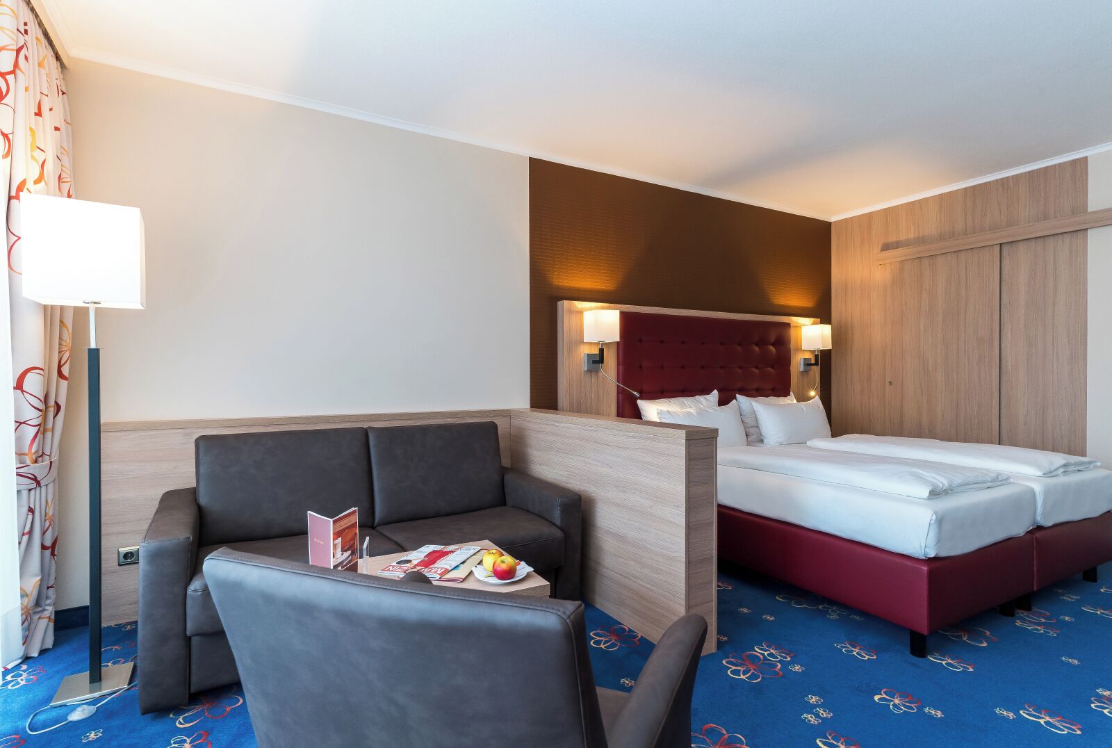 Une chambre d’hôtel avec un lit, un canapé, une table avec un magazine et des fruits, et une lampe.