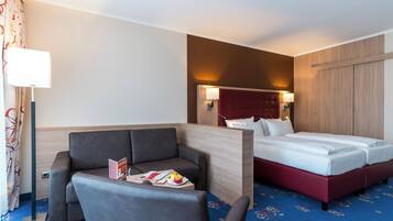 Une chambre d’hôtel avec un lit, un canapé, une table avec un magazine et des fruits, et une lampe.