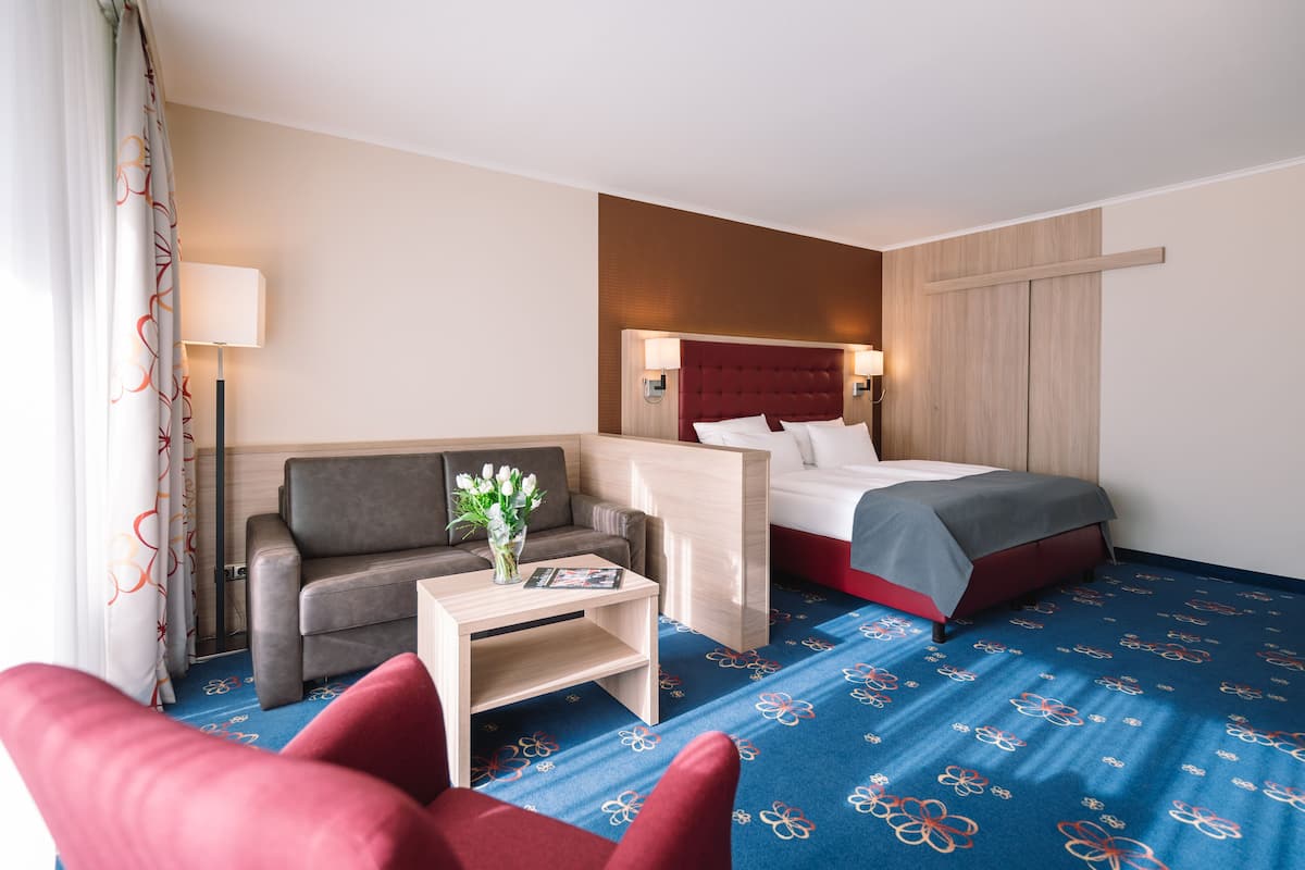 A hotel room with a bed, a sofa, a small table with a vase of flowers, and a red armchair.