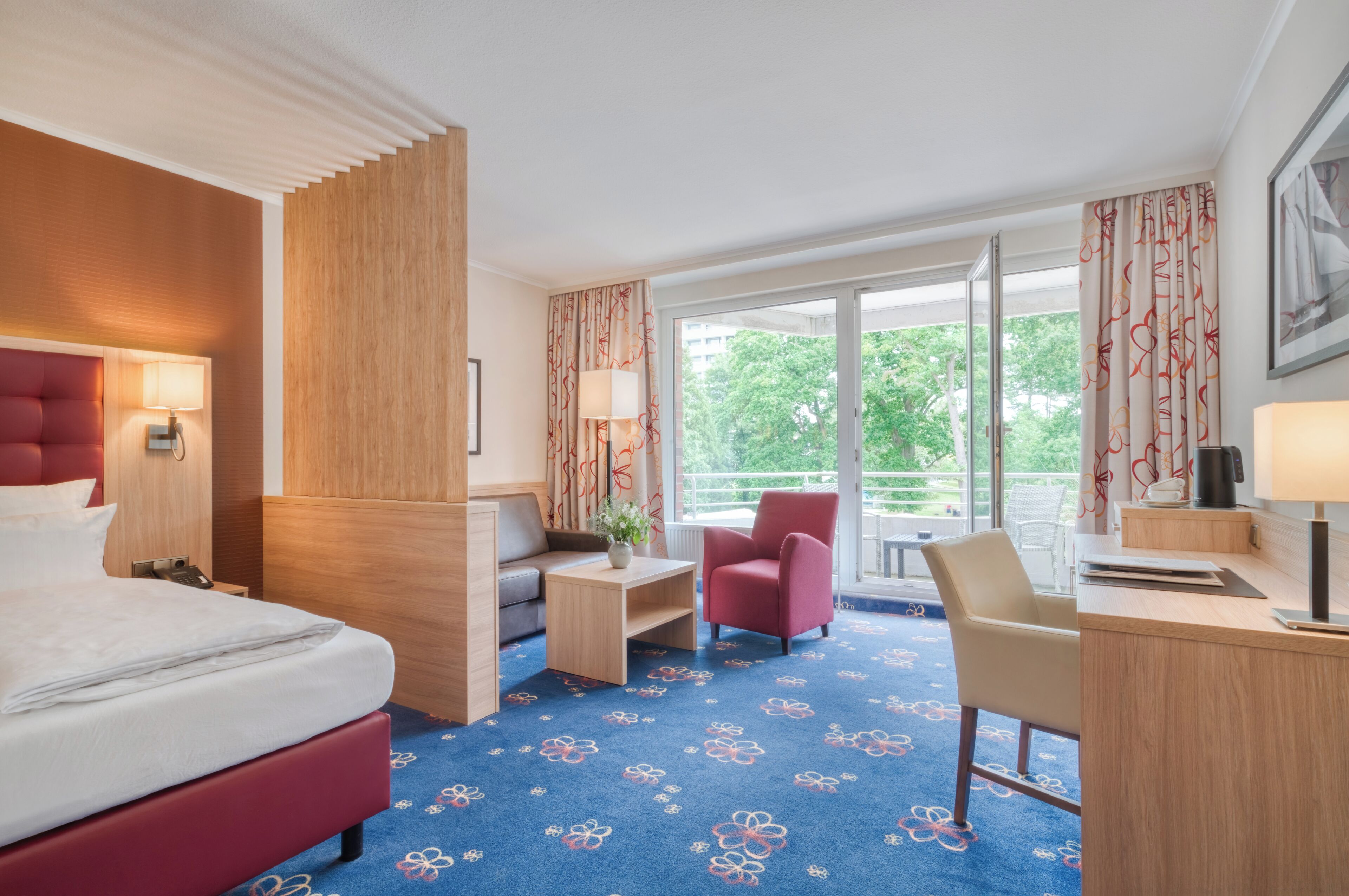 Una camera d'albergo con un letto, una scrivania, una sedia, un divano e un balcone con vista sugli alberi.