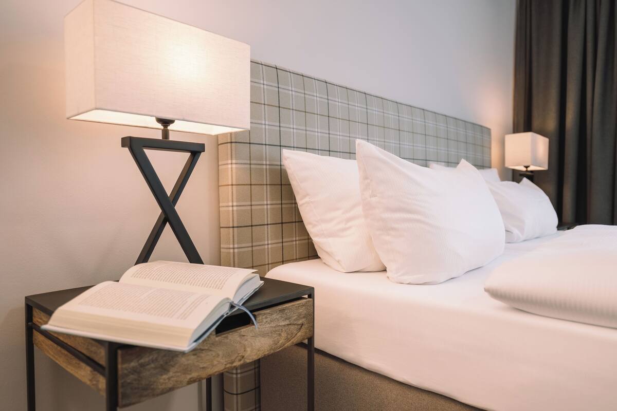 A hotel room with a bed, two bedside lamps, a nightstand with an open book, and a checkered headboard.
