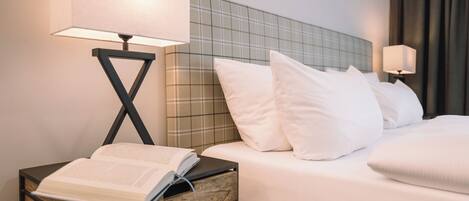 A hotel room with a bed, two bedside lamps, a nightstand with an open book, and a checkered headboard.