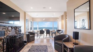 A modern living room with a stone fireplace, a large flat-screen TV, comfortable seating, a dining area with a view of the ocean, and a large mirror on the wall. - Coastal Retreat Waterfront, Deck, Awesome Views (Wading River)