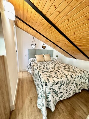 A bedroom with a sloped ceiling, a bed with a floral bedspread, a grey headboard, and a hanging heart-shaped decoration. - Casa Secunda Romar (Córdoba)