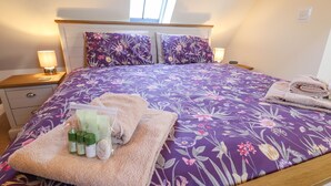A neatly made bed with floral bedding, a bedside table with a lamp, and a small bedside table with a phone. - Saddle Rack Cottage (Louth)