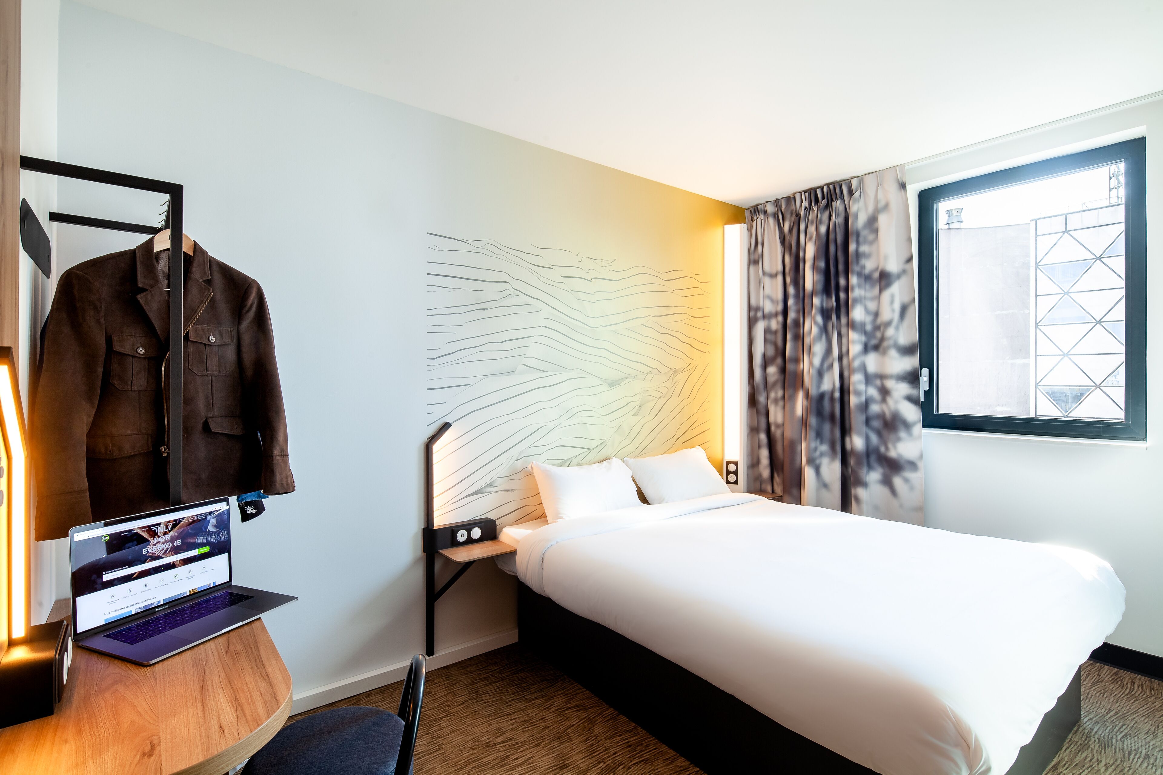 Habitación de hotel con una cama, un escritorio con una computadora portátil, un perchero y una pared con diseño geométrico.
