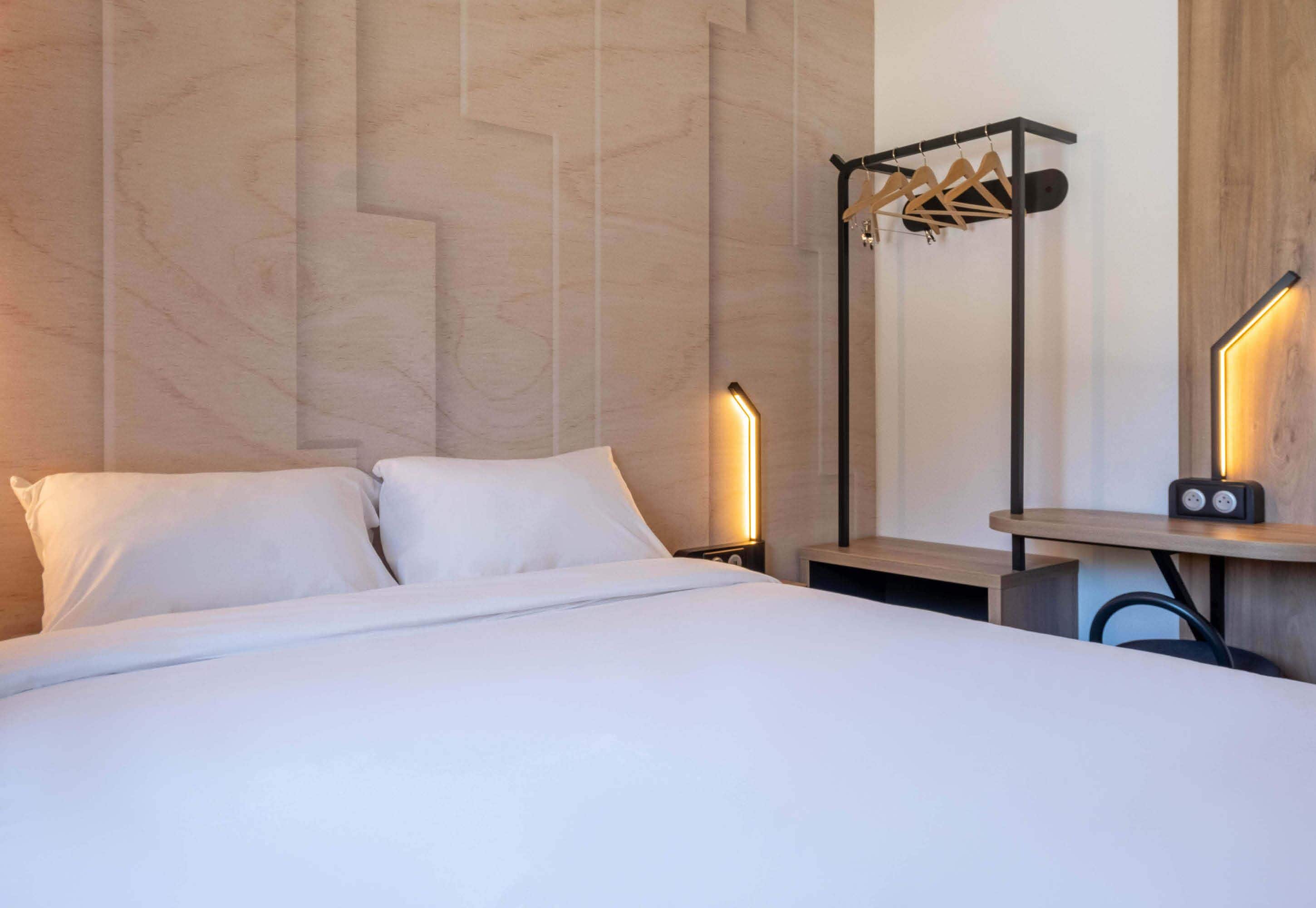 Ein Hotelzimmer mit einem Bett, einem Nachttisch, einer Wandlampe, einem Holzbodenregal und einem Kleiderhaken.