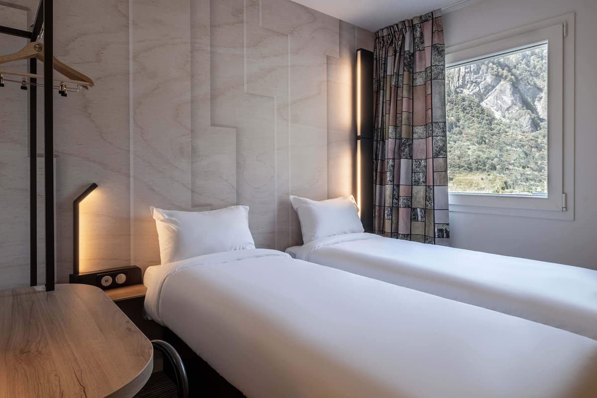 Habitación de hotel con dos camas, una mesa de madera, una mesita de noche con lámpara y ventana con vista a montañas.