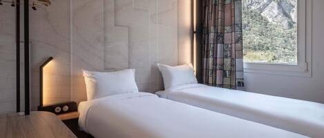 A hotel room with two beds, a wooden table, a nightstand with a lamp, and a window with a view of mountains.