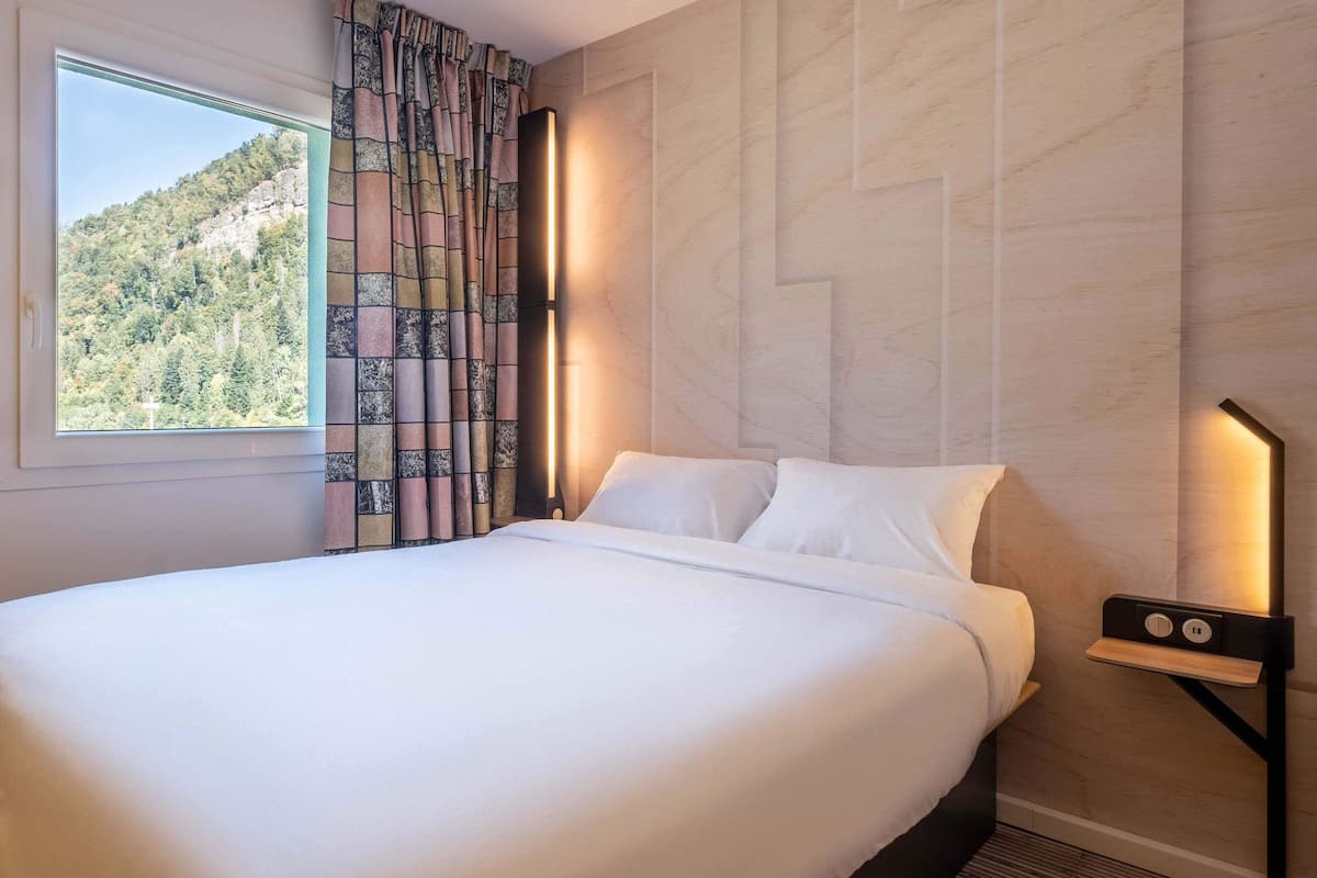 Una cama bien hecha con sábanas blancas, una ventana con vistas a una ladera boscosa y una mesita de noche fijada a la pared con una lámpara.