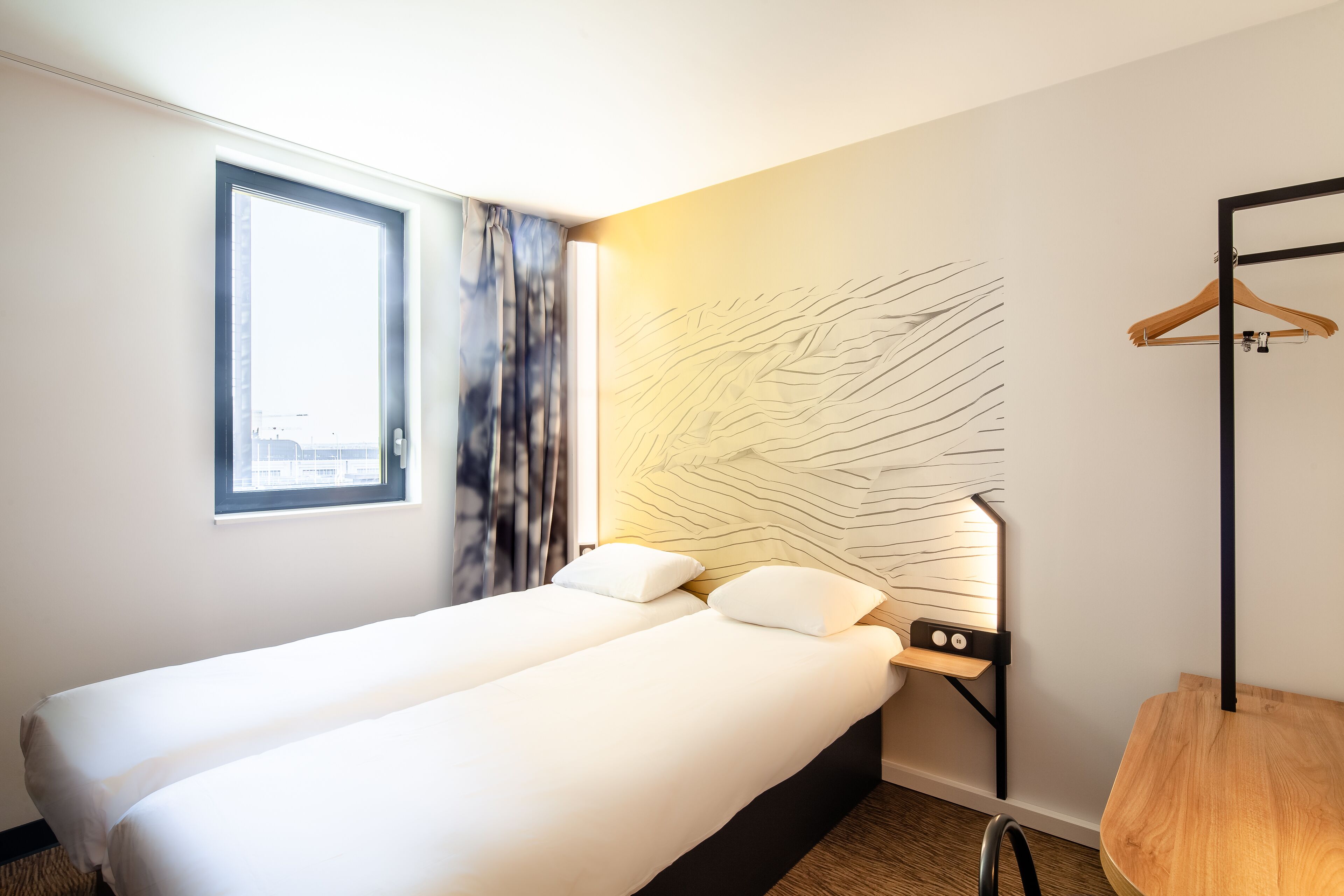 A hotel room with a bed, a window, a wall art piece, and a wooden chair.