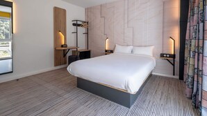 Una habitación de hotel moderna con una cama grande, un escritorio con silla y una mesita de noche con lámpara.