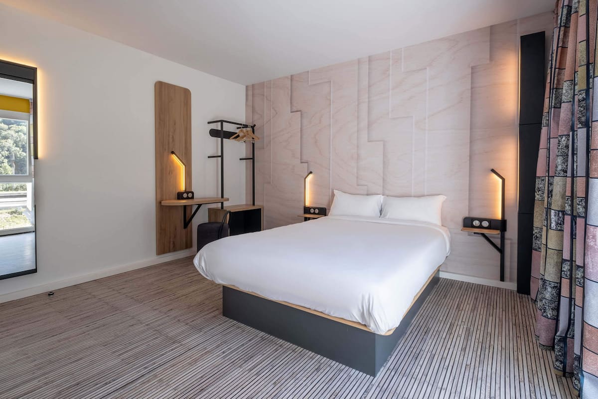 Una habitación de hotel moderna con una cama grande, un escritorio con silla y una mesita de noche con lámpara.