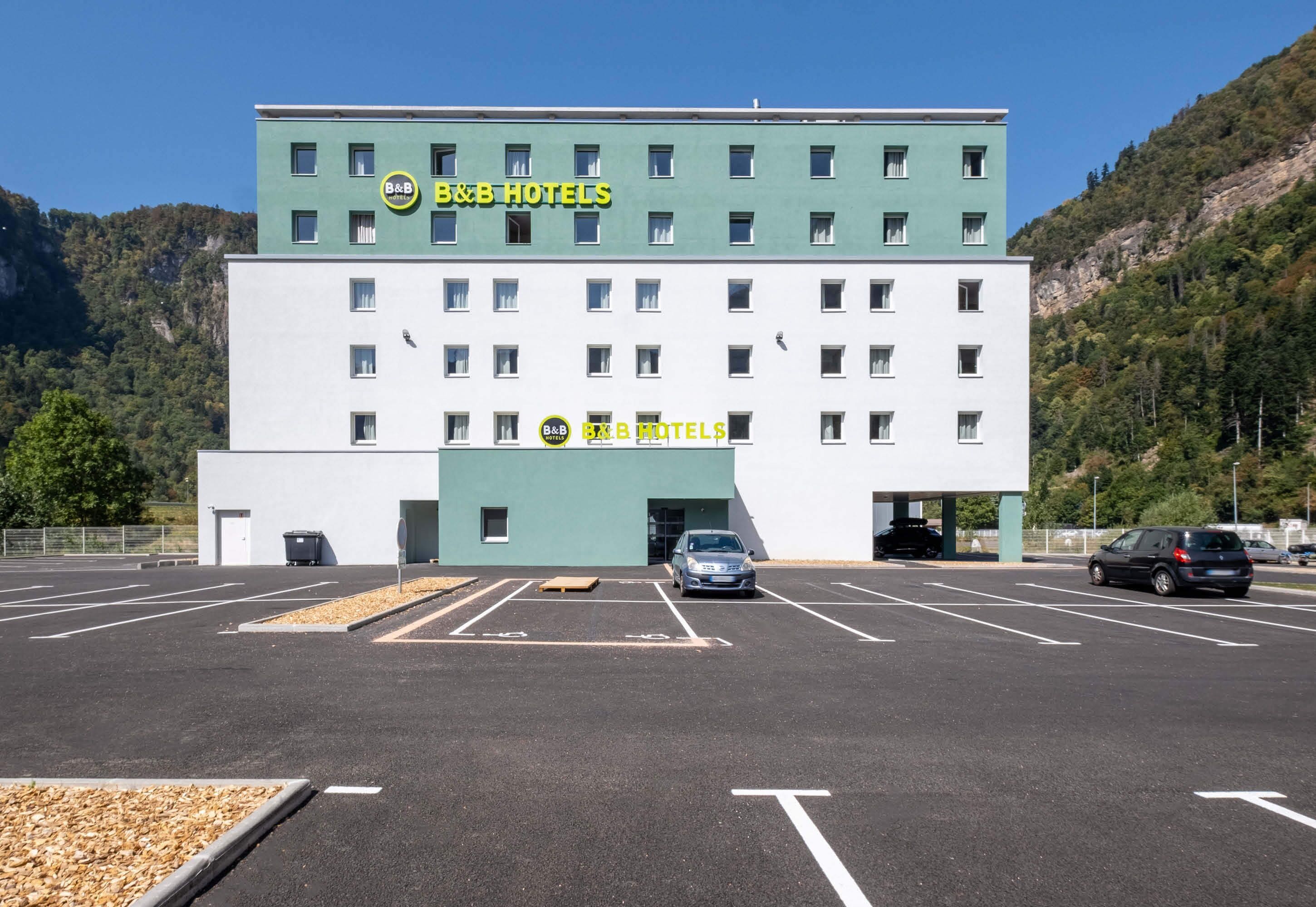 Un hotel tipo casa de huéspedes con fachada verde, varias ventanas y un estacionamiento al frente.