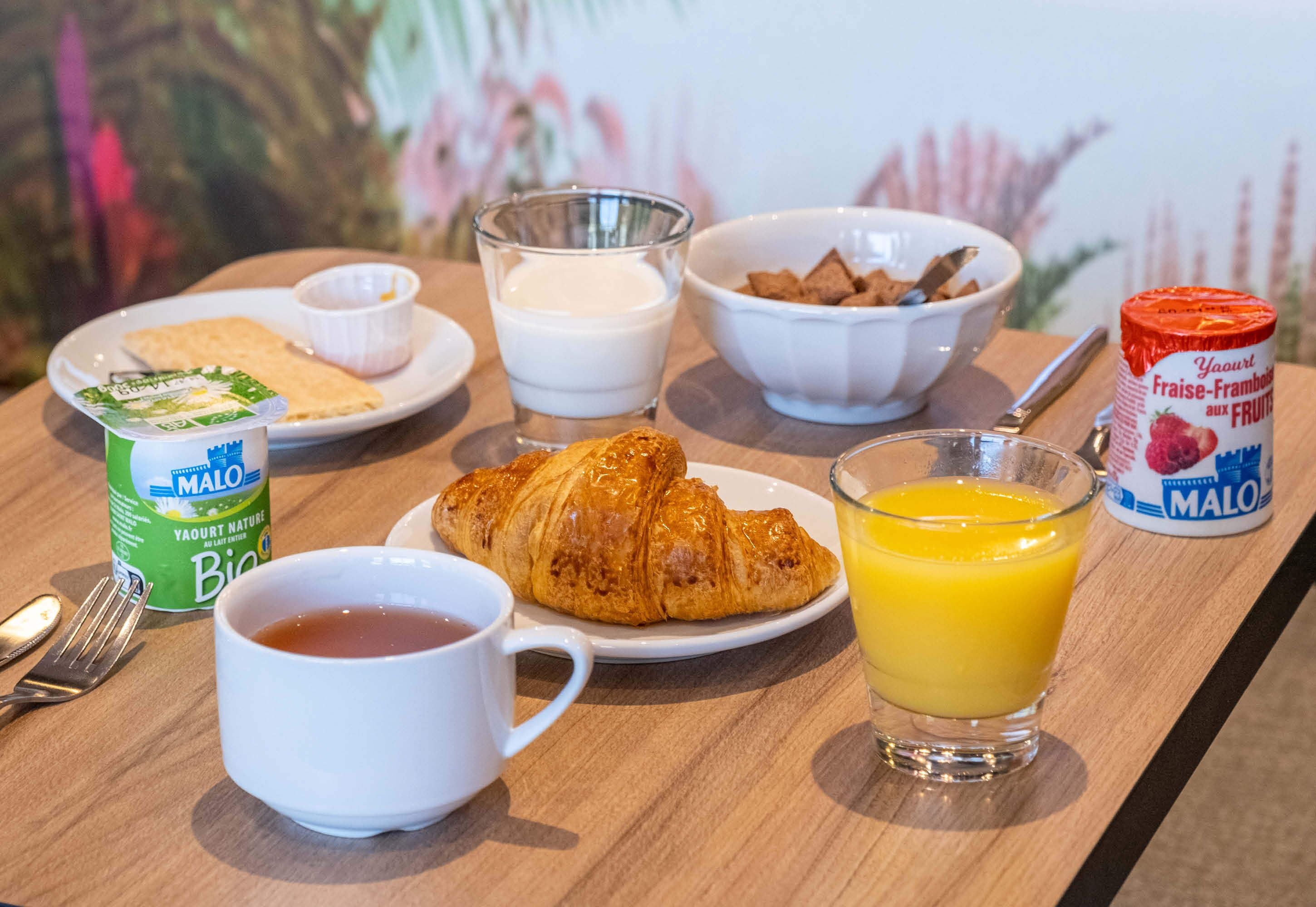Ein Frühstückstisch mit Croissants, Orangensaft, einer Tasse Tee, einer Schüssel Müsli und einem Joghurtbecher.